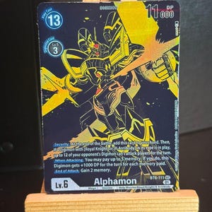 Peut inclure: Une carte à collectionner Digimon représentant Alphamon, un Digimon de niveau Méga. La carte a un fond noir avec des accents et du texte dorés. La carte affiche les statistiques, les capacités et le texte "Alphamon". La carte est présentée sur un petit chevalet en bois.