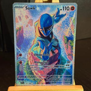May include: A holographic Sawk Pokémon trading card. The card features a blue character with the text "Sawk" at the top. The card also includes the text "Elbow Strike" and "Rising Chop". The card is displayed on a small wooden easel.