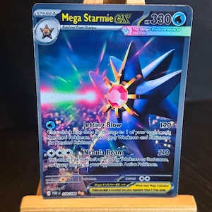 Puede incluir: Una carta coleccionable de Pokémon Mega Starmie EX. La carta presenta una criatura en forma de estrella azul y rosa con el texto "Mega Starmie EX", "Jetting Blow" y "Nebula Beam". La carta se muestra en un pequeño caballete de madera.