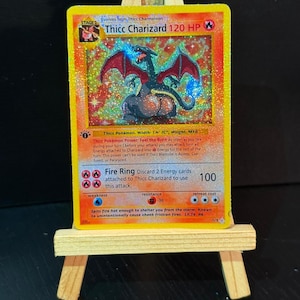 May include: A holographic Thicc Charizard Pokémon card, displayed on a small wooden easel. The card features a red and black dragon illustration with the text "Thicc Charizard 120 HP". The card is a Stage 2 evolution.