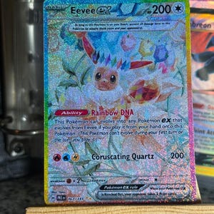 Puede incluir: Una carta coleccionable holográfica de Eevee ex con 200 HP, que presenta una corona de cristal Tera y la habilidad Rainbow DNA. La carta muestra el ataque Coruscating Quartz, una debilidad a los tipos lucha y el número 167/131 con acabado arcoíris.