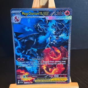 May include: A holographic Mega Charizard X EX trading card. The card features a blue and red illustration of Charizard with the text "Mega Charizard X EX" and "Inferno X". The card is displayed on a small wooden easel.
