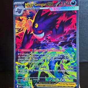 Könnte beinhalten: Eine holografische Mega Gengar EX Pokémon-Sammelkarte. Die Karte zeigt ein lila Gengar mit einem finsteren Grinsen, vor einem feurigen roten und gelben Hintergrund. Der Kartentext enthält "Shadowy Concealment" und "Void Gale".