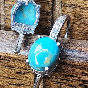 May include: A pair of silver-toned earrings featuring oval-shaped turquoise cabochons with visible yellow matrix. Each earring has a curved hook with a row of small, clear, round-cut stones set along the front edge and four prongs holding the stone.