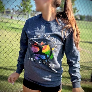 May include: A dark gray long-sleeve shirt with the text "THROW SPEARS NOT SHADE" and a colorful graphic of a person throwing a spear. The shirt is paired with black shorts. The image is taken outdoors.