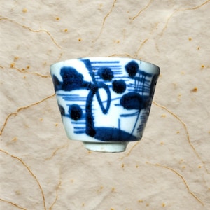 Op de afbeelding: Een kleine, witte keramische kom met een blauw bloemmotief. De kom heeft een uitlopende vorm en een kleine voet. Het blauwe ontwerp bevat abstracte bloem- en geometrische vormen. De kom is waarschijnlijk voor het serveren van thee of kleine porties.