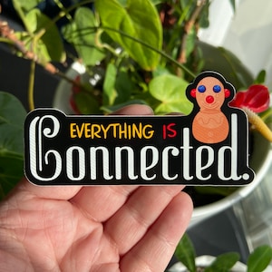 May include: A black and white sticker with the text "Everything is connected." in yellow and white. A cartoon gingerbread man with blue eyes and red cheeks is on the right side of the sticker.