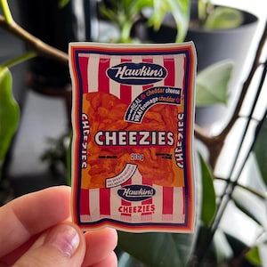May include: A packet of Hawkins Cheezies, with a red and white striped design. The package displays the brand name "Hawkins" and the product name "Cheezies". The text "REAL cheddar cheese" is also visible, along with the weight of the contents, 210g.