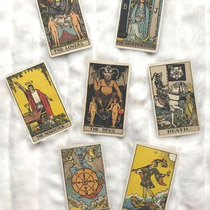 May include: A collection of vintage tarot cards featuring illustrations and titles such as "The Lovers," "The High Priestess," "The Magician," "The Devil," "Death," "Wheel of Fortune," and "The Fool." The cards have a cream-colored border and are arranged on a white surface.