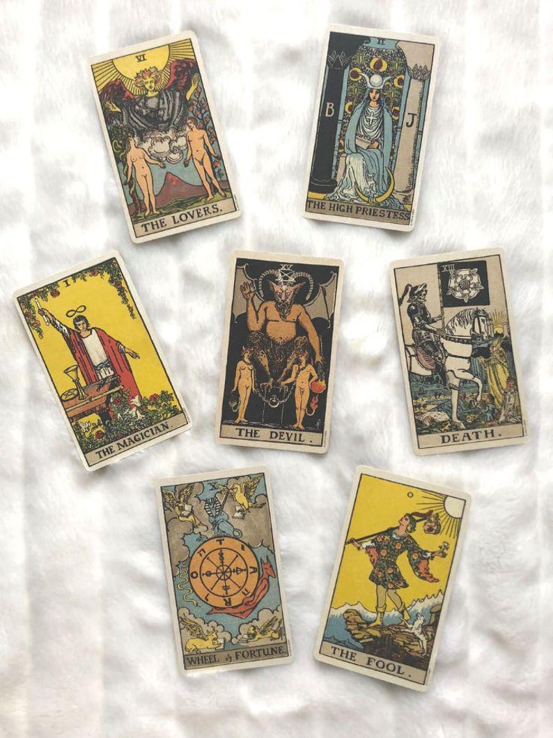 May include: A collection of vintage tarot cards featuring illustrations and titles such as "The Lovers," "The High Priestess," "The Magician," "The Devil," "Death," "Wheel of Fortune," and "The Fool." The cards have a cream-colored border and are arranged on a white surface.