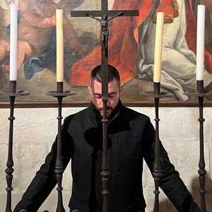 May include: A person stands before a wrought iron cross with a crucifix, flanked by tall candles in ornate holders. The background features a religious painting with warm colors, creating a solemn atmosphere. The person is wearing a black jacket.