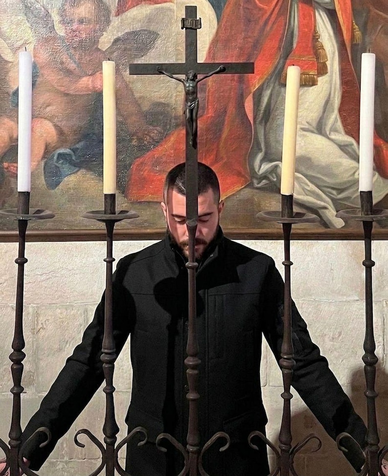 May include: A person stands before a wrought iron cross with a crucifix, flanked by tall candles in ornate holders. The background features a religious painting with warm colors, creating a solemn atmosphere. The person is wearing a black jacket.