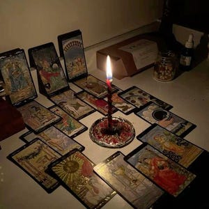 May include: A lit red candle sits on a decorative plate surrounded by tarot cards. The cards are arranged on a white surface. A box and a jar of nuts are in the background. The scene is dimly lit.