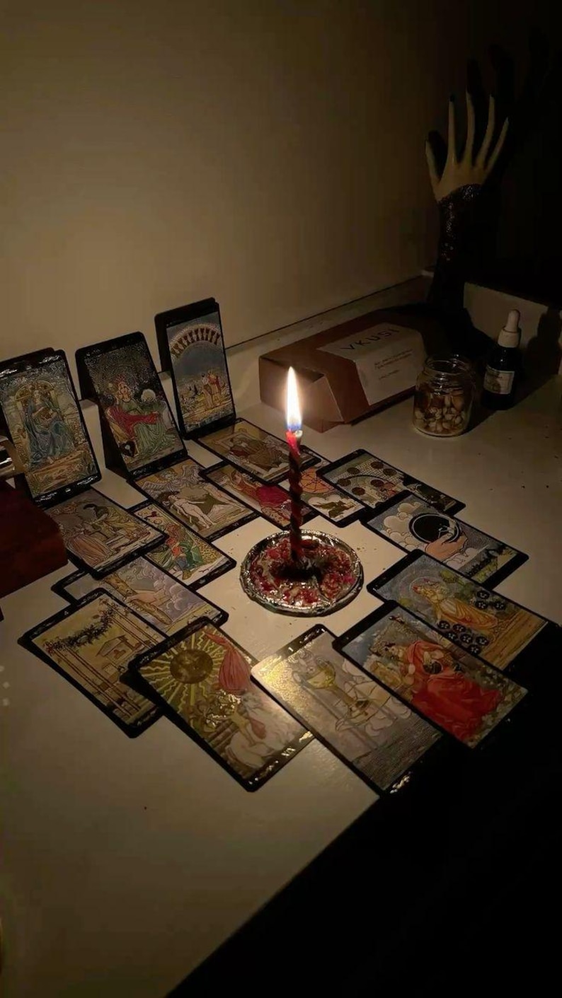 May include: A lit red candle sits on a decorative plate surrounded by tarot cards. The cards are arranged on a white surface. A box and a jar of nuts are in the background. The scene is dimly lit.