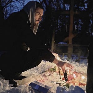 May include: A person in a hooded sweatshirt kneels in the snow, reaching towards lit candles and flowers. A wooden cross stands in the background, with bare trees and a dark sky.