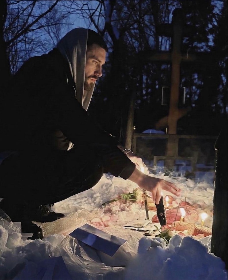 May include: A person in a hooded sweatshirt kneels in the snow, reaching towards lit candles and flowers. A wooden cross stands in the background, with bare trees and a dark sky.
