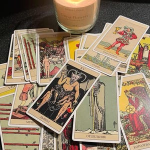 May include: A lit candle in a glass jar with the text "Daisy Flowers" sits near a collection of tarot cards. The cards feature illustrations and text in various languages, including "Дьявол" and "ОТШЕЛЬНИК".