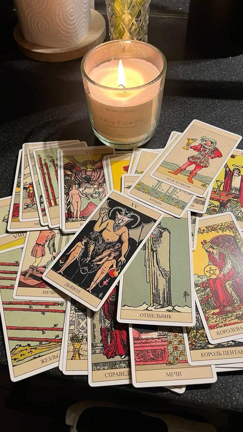 May include: A lit candle in a glass jar with the text "Daisy Flowers" sits near a collection of tarot cards. The cards feature illustrations and text in various languages, including "Дьявол" and "ОТШЕЛЬНИК".