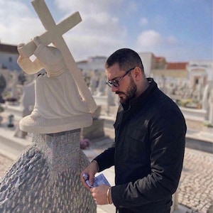 May include: A white stone sculpture of a figure carrying a cross. The sculpture is set on a textured base. A person wearing glasses and a black jacket is standing nearby, holding money. The background shows a cemetery under a blue sky.