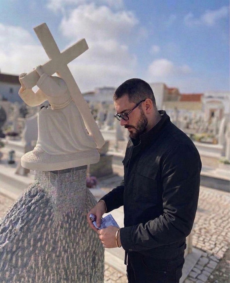 May include: A white stone sculpture of a figure carrying a cross. The sculpture is set on a textured base. A person wearing glasses and a black jacket is standing nearby, holding money. The background shows a cemetery under a blue sky.