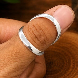 May include: A smooth, polished silver band ring with a rounded profile. The ring features a simple, continuous design with a reflective metallic surface. The band is shown encircling a thumb, highlighting its plain, unadorned, and circular shape.