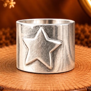 May include: A wide silver-toned ring featuring a raised five-pointed star design on its front surface. The ring has a smooth, polished finish and a cylindrical band shape, showcasing a clean and simple aesthetic without any additional embellishments.