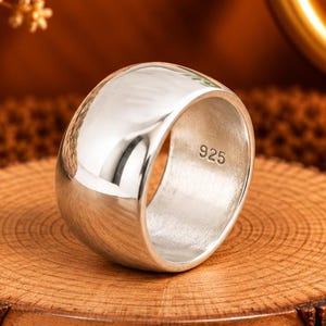 May include: A wide, polished silver band ring with a smooth, reflective surface. The interior of the ring is clearly stamped with the hallmark 925, indicating sterling silver composition. The ring is positioned on a circular wooden display base.