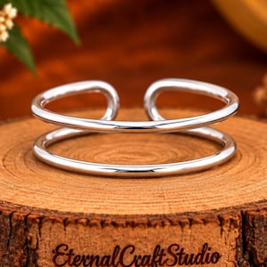 Può includere: Un bracciale a fascia aperta color argento lucido con un design a doppio cerchio. Il bracciale è posizionato su una fetta di legno circolare che presenta il testo EternalCraftStudio e un simbolo a forma di cuore incisi sul davanti.