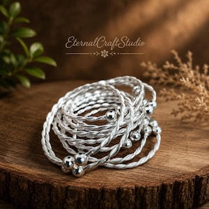 Può includere: Un set di anelli in filo ritorto color argento, ciascuno caratterizzato da una piccola perla sferica alle estremità. Gli anelli sono impilati in una disposizione circolare. Il testo EternalCraftStudio è visibile nella parte superiore dell'immagine.
