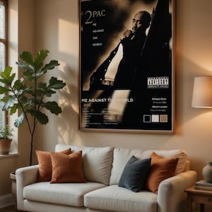 May include: A framed poster featuring 2Pac's "Me Against the World" album cover hangs above a cream-colored sofa with several throw pillows. The poster is in black and white, with the album title and artist's name prominently displayed. The room has a warm, inviting atmosphere.