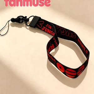 May include: Black lanyard with red text that reads "JIN JUNG" and "SUGA". The lanyard has a black clasp and a small loop. The word "fanmuse" is written in pink above the lanyard.