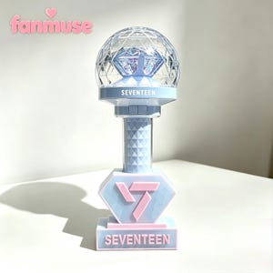 May include: A light blue and pink concert light stick with the word "SEVENTEEN" printed on the base and the top. The light stick has a diamond-shaped head with a geometric pattern and a clear, faceted globe on top.