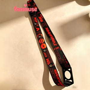 May include: Black lanyard with red text that reads "JUNGKOOK" and other names. The lanyard has a black plastic clasp and metal eyelets. The word "fanmuse" is in pink text in the upper left corner.
