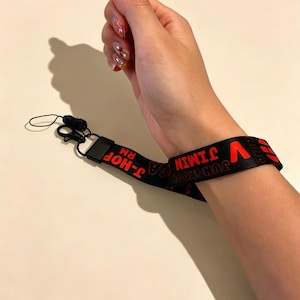 May include: Black wrist lanyard with red text featuring the names of band members. The lanyard has a black clasp and a black cord. The word "fanmuse" is in pink above the lanyard.
