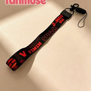 May include: Black lanyard with red text featuring the names of band members. The lanyard has a black clip and a small black loop. The word "fanmuse" is in pink script at the top.