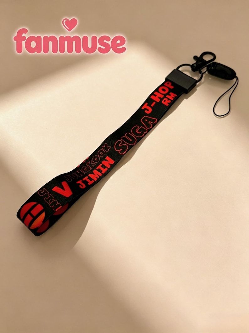 May include: Black lanyard with red text featuring the names of band members. The lanyard has a black clip and a small black loop. The word "fanmuse" is in pink script at the top.