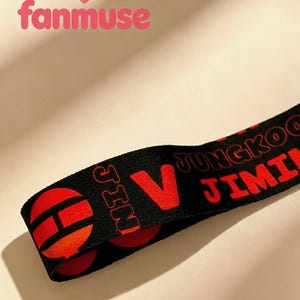 May include: Black lanyard with red text and design elements. The text includes the names "JUNGKOO", "JIMIN", and "JIN", along with a large red "V". The lanyard also features red circular designs.