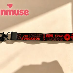 May include: Black lanyard with red text that reads "JIN JIMIN JUNGKOOK RM SUGA J-HOPE". The lanyard has a black clasp and a small black loop. The word "fanmuse" is in pink above the lanyard.