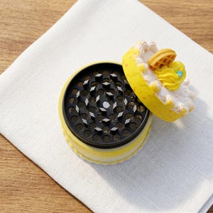 Op de afbeelding: Een gele en zwarte kruidenmolen met een decoratief cakevormig deksel. Het deksel heeft witte glazuur, gele glazuur en een kleine wafel. De molen is open en toont de maaltanden.