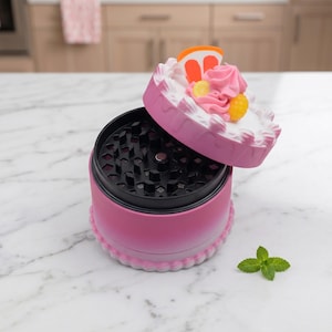 Op de afbeelding: Een roze kruidenmolen met een cake-thema deksel. Het deksel is versierd met witte glazuur, roze glazuurkrullen en oranje en gele fruitschijfjes. De behuizing van de molen heeft een roze verloop en de basis heeft een decoratieve witte rand.