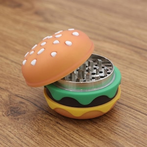 May include: A hamburger-shaped herb grinder with a silicone exterior designed to look like a burger. The top bun is light orange with white sesame seed details. The base features layers of green lettuce, dark brown meat, and yellow cheese.