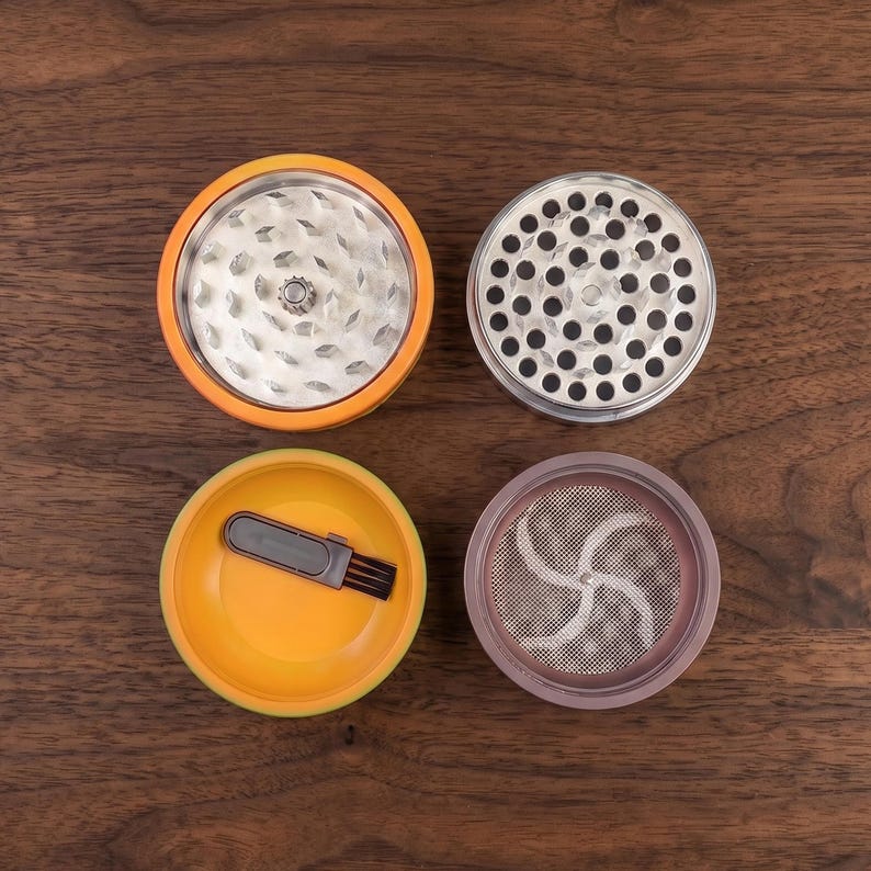 May include: A four-piece herb grinder set shown in a disassembled state. The set includes an orange-rimmed grinding chamber with metal teeth, a silver-toned lid with circular holes, an orange base, and a silver-toned mesh screen with a small brush.