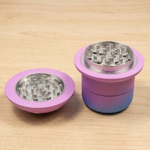 Puede incluir: Un molinillo de hierbas de dos piezas con un acabado degradado que va del rosa al azul. La sección superior está separada y colocada junto al cuerpo principal, revelando dientes metálicos plateados diseñados para moler en ambas superficies.
