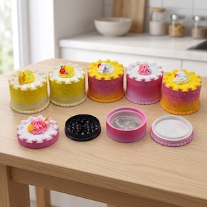 May include: A collection of novelty grinders designed to resemble miniature cakes. Each grinder features a cylindrical body with a decorative top, mimicking frosting and toppings. The colors include yellow, pink, and white, with a black grinder component visible.