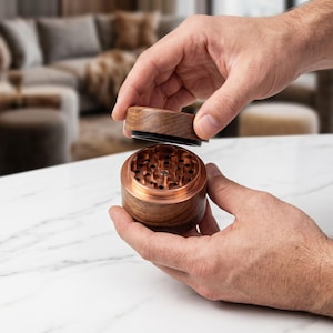 May include: A cylindrical herb grinder featuring a dark brown wood exterior and a metallic copper-coloured interior. The device consists of two parts, with the top lid removed to reveal sharp, diamond-shaped teeth designed for grinding herbs.