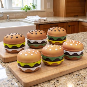 May include: Six round, burger-shaped containers with lids, each featuring a tan bun top with black or white sesame seed details. The containers have layered, wavy-edged sections in shades of brown, dark brown, light green, and yellow.