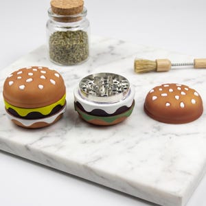 May include: A three-piece herb grinder shaped like a hamburger with a brown bun top, a green lettuce layer, and a dark brown patty layer. The center section reveals a metallic grinding chamber with sharp teeth, accompanied by a small cleaning brush.