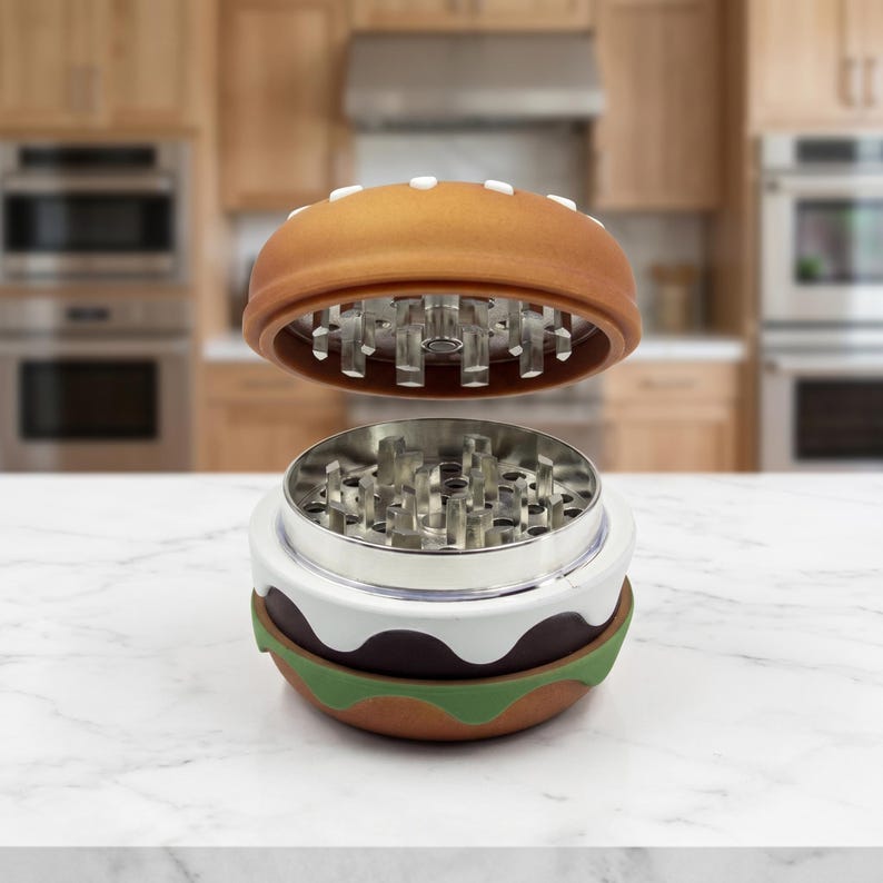 May include: A multi-piece herb grinder shaped like a hamburger. The top lid is brown with white sesame seed details. The interior features sharp metal teeth for grinding. The base layers are colored white, dark brown, and green to resemble a bun.