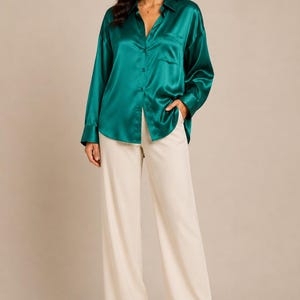 May include: A woman wearing a teal green satin button-down shirt with a chest pocket and cream-colored wide-leg pants. The shirt is buttoned at the center and the pants are high-waisted.