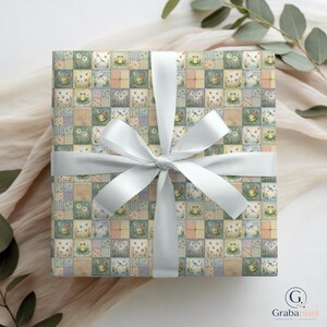 K&ouml;nnte beinhalten: Ein quadratisches Geschenk, verpackt in gemustertem Papier mit einem wei&szlig;en Satinband, das zu einer Schleife gebunden ist. Das Papier zeigt ein Patchwork-Design mit Illustrationen von Fr&ouml;schen, Libellen und Blumen in Pastellfarben. Die Schleife ist mittig auf dem Geschenk.